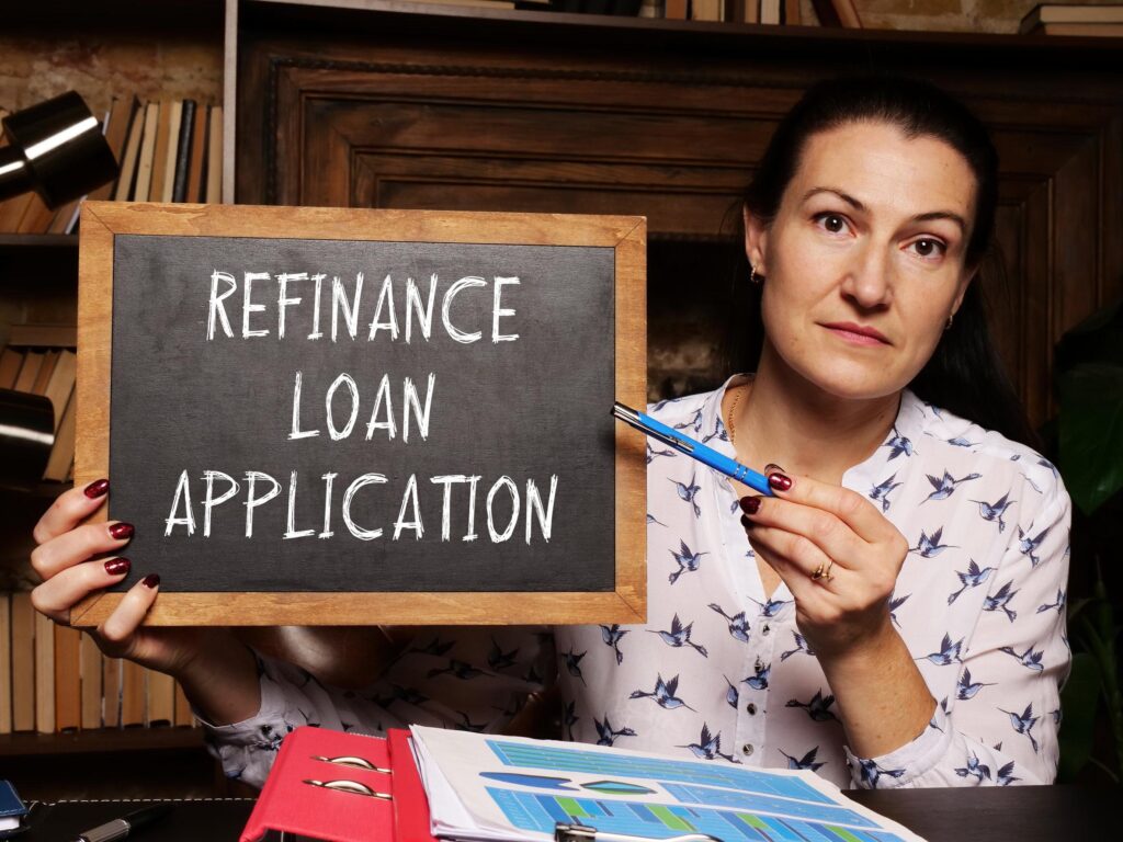 Lady pointing with a pen at a “Refinance Loan Application” signboard she’s holding.
