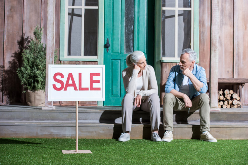 Upset senior couple sitting on the porch of their home with a sale sign.
